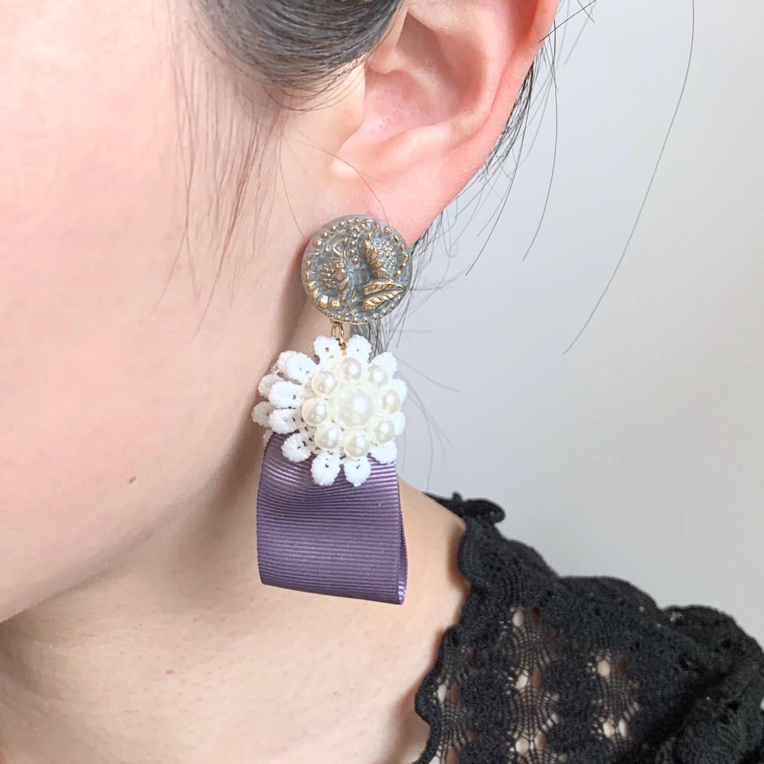 ラベンダー色の大ぶりポンポンとフェイスモチーフピアス ラベンダー色のリボンとお花モチーフの〈ピアス/イヤリング〉 | Dua