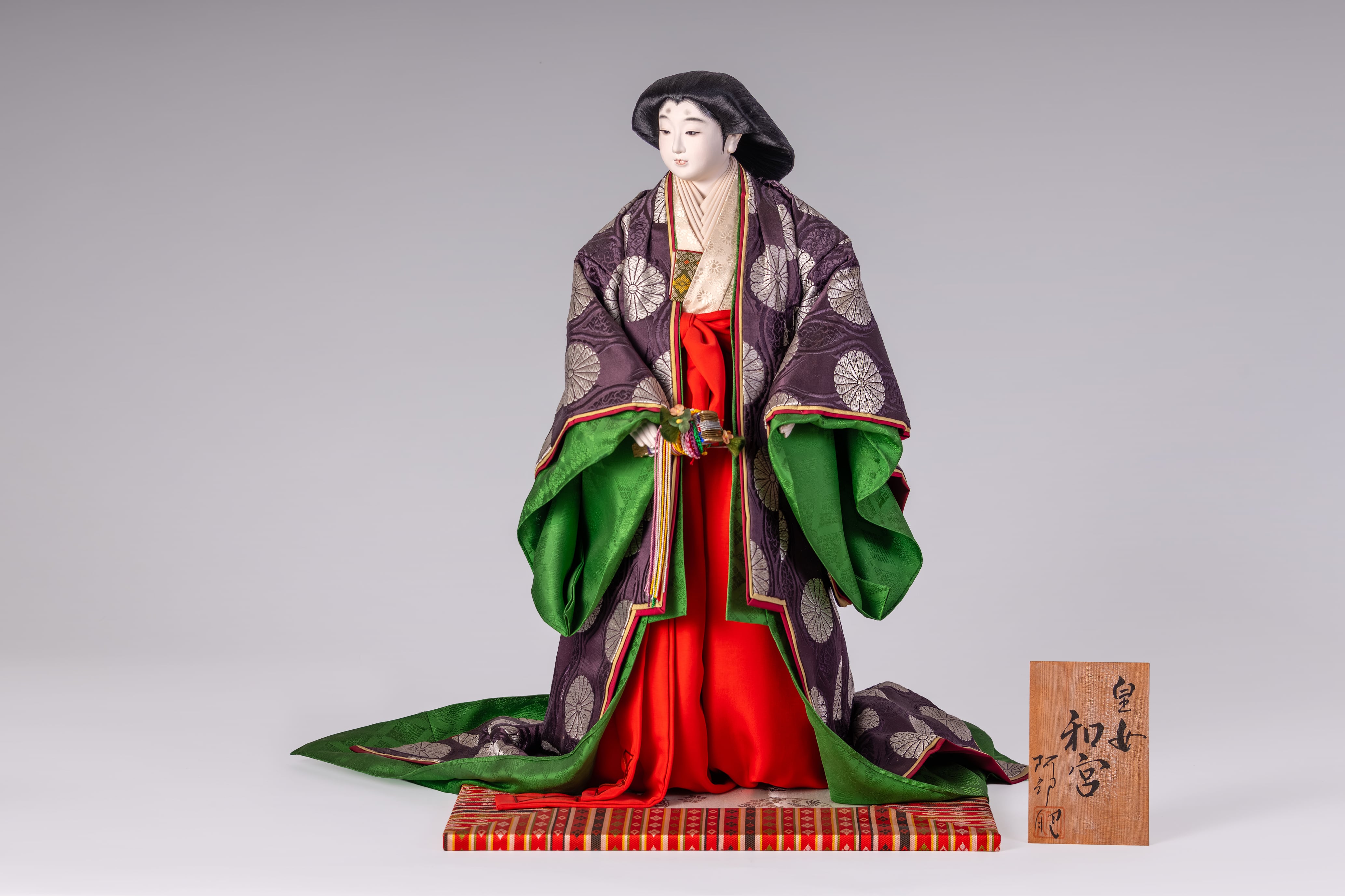 人形作品「皇女 和宮」は、菊花文様の小袿をまとい、気品と静かな格調を美しく表現した一点ものの作品です。

深い紫を基調とした装束に、緑や朱の重なりが華やかさを添え、凛とした表情や美しい後ろ姿まで丁寧に仕立てられています。日本の伝統美と歴史の趣を感じさせる、鑑賞用にも空間演出にもふさわしい上品な一作です。