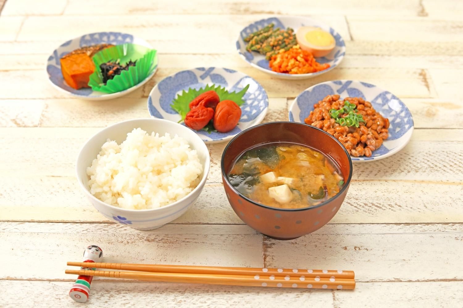 白木の台に並んだ和食。白いごはん、わかめと豆腐の味噌汁、梅干し、納豆、煮物など