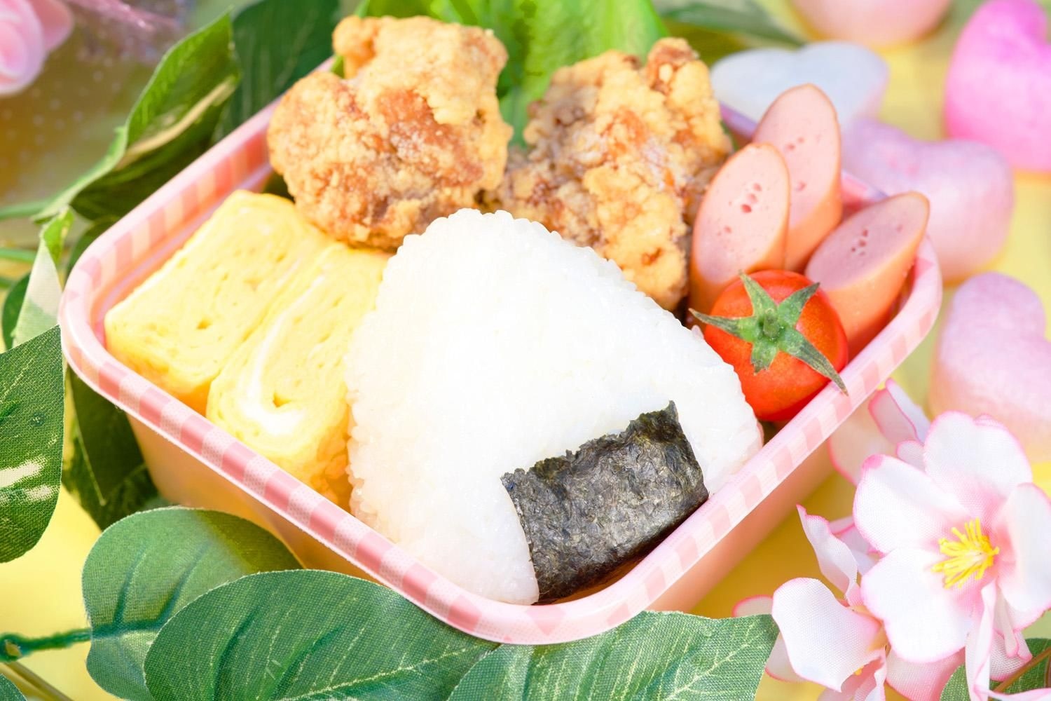 お弁当箱に、卵焼きの黄色、ミニトマトの赤、からあげやおにぎりを詰めた彩りのよいお弁当の一例