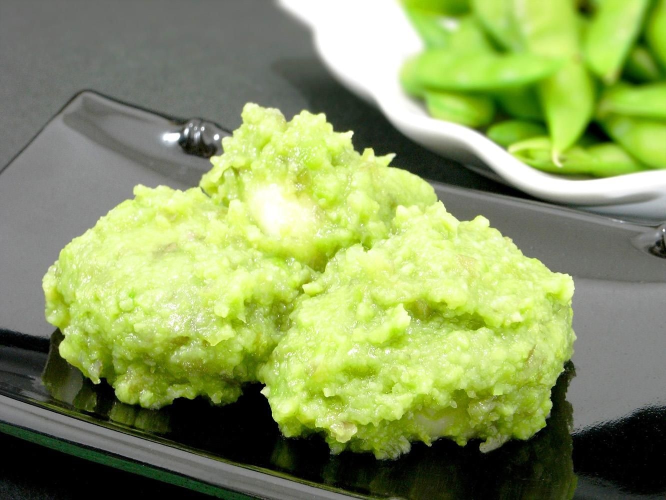 黒いお皿にのせた、枝豆をすりつぶした緑のあん（ずんだ）。奥にさや付きの枝豆