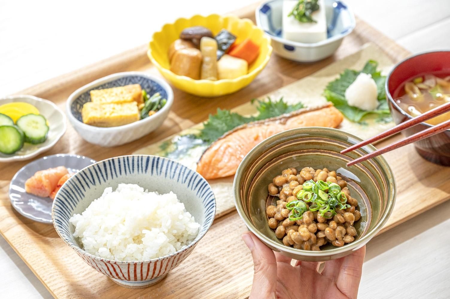 木のトレイに並ぶ朝の和定食。白いごはん、納豆、玉子焼き、焼き鮭、煮物、豆腐、お味噌汁、きゅうりの浅漬け